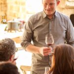 A host presenting a glass of red wine to seated guests in the Villa
