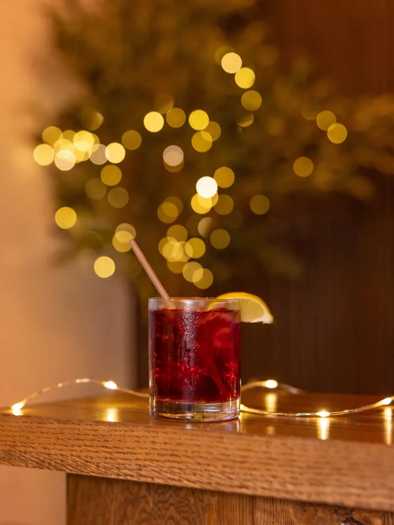 Sunstone ELIXIR built as a mocktail with lemon and a straw, on a wooden bar beside a string of fairy lights