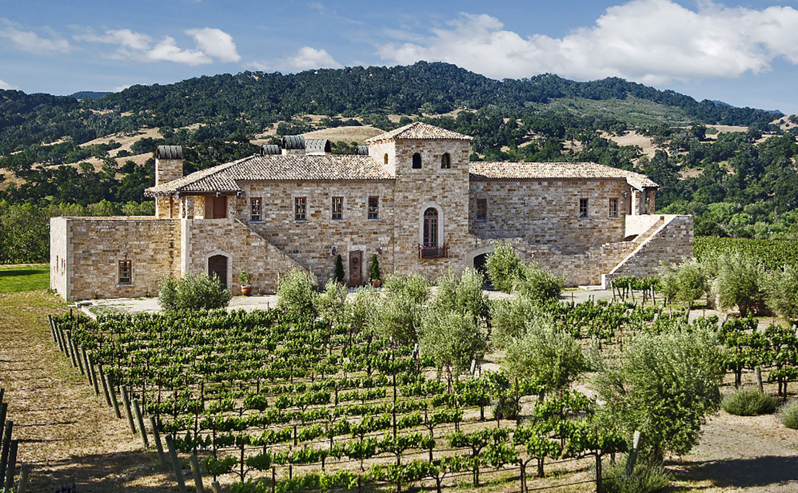 The Villa, a two-storey limestone building with a tiled roof and square tower, seen across its vineyard with wooded hills behind.
