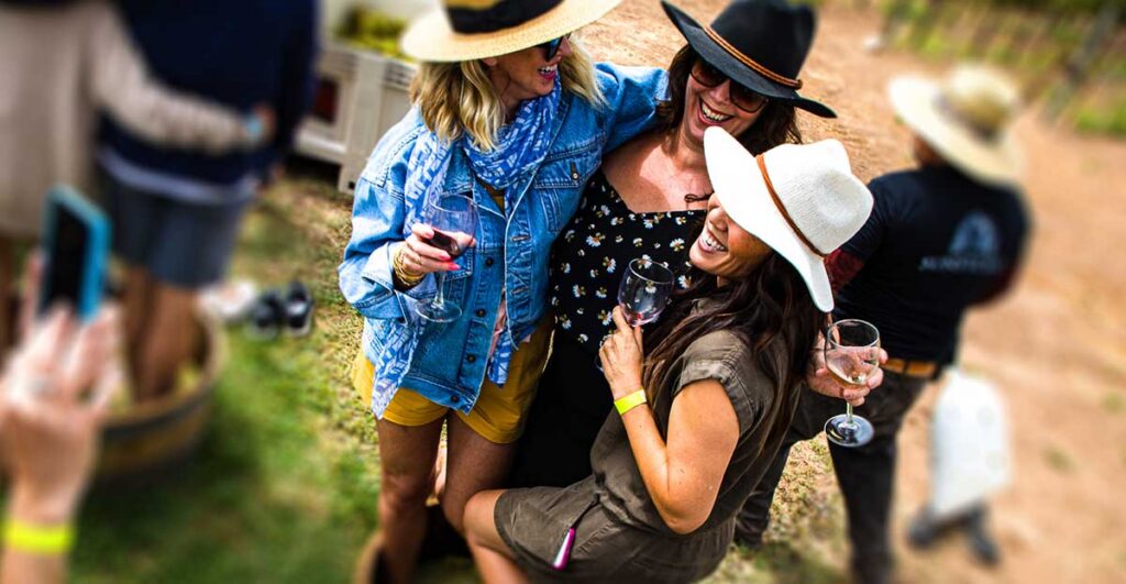 Three guests in sun hats laughing together with glasses of wine beside a stomping barrel